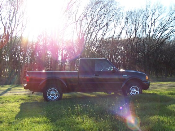 Side view of a dark-colored Ford Ranger pickup truck parked in a grassy field with trees in the background. Sun flares are visible