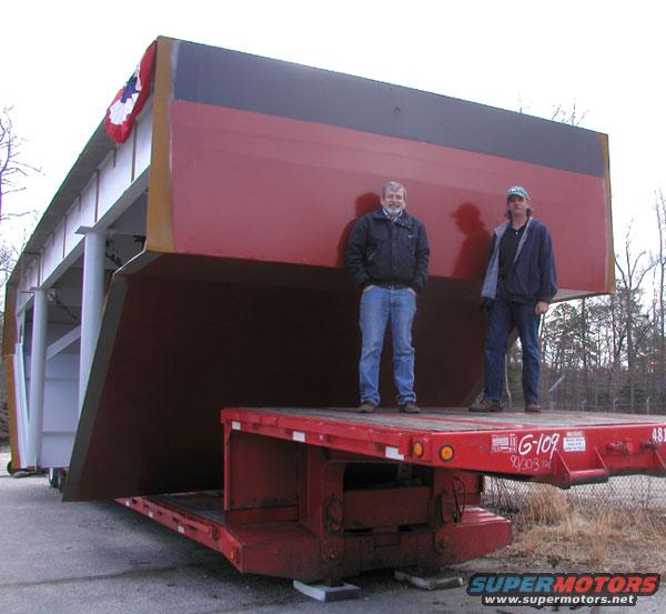 Two men stand on a flatbed trailer next to a massive red and black cargo container with a small American flag decoration