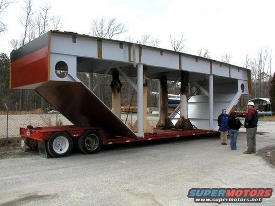 Red trailer carrying a large, gray and red industrial structure with people standing nearby