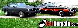 Two 1978 Ford Mustang IIs parked side-by-side, one matte black, the other black with red 'Cobra' graphics