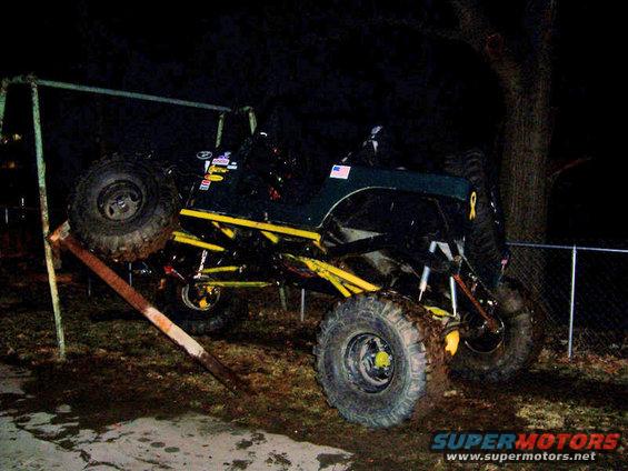 Modified Jeep with large tires stuck on a metal bar, next to a tree and fence