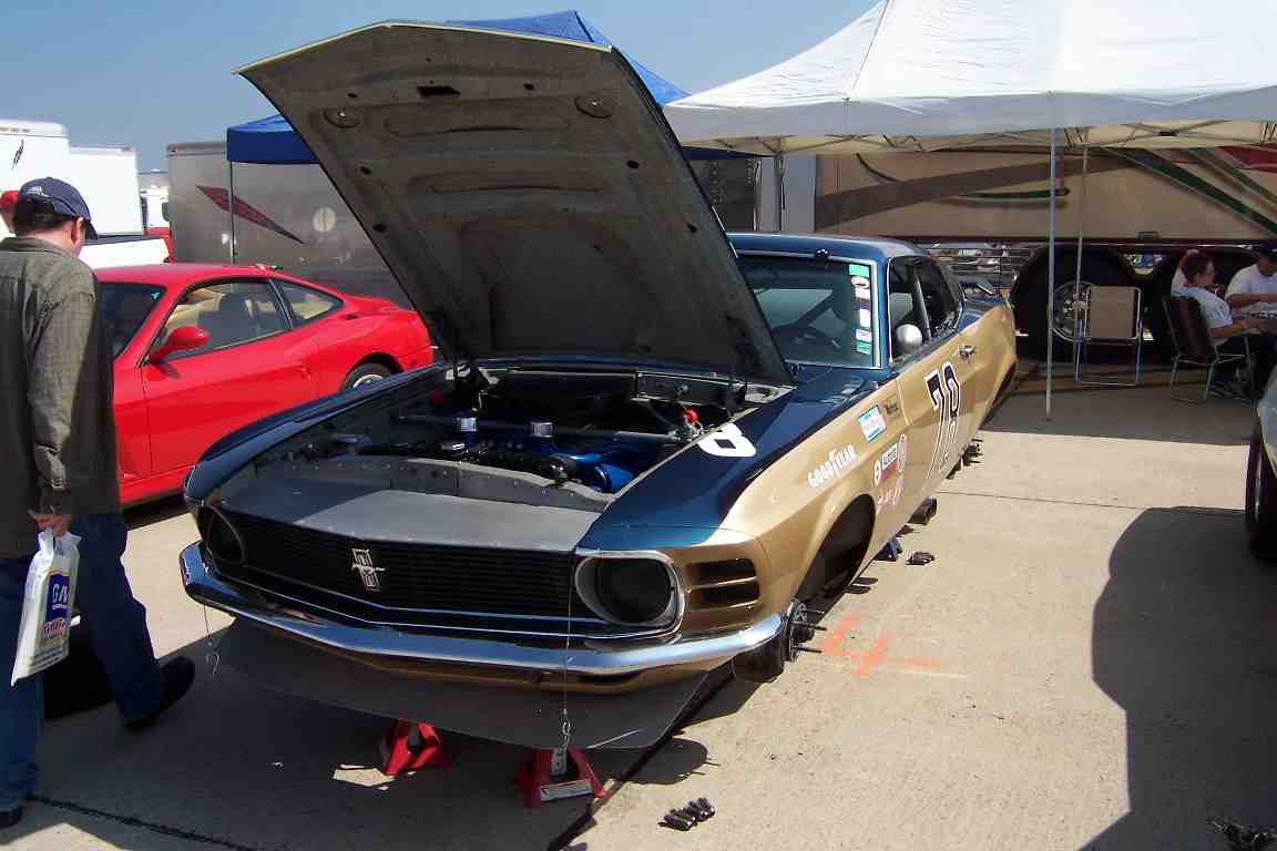 1970 Ford Mustang race car with hood open, jack stands under front, Goodyear tires, number 70 on door. Red car in background