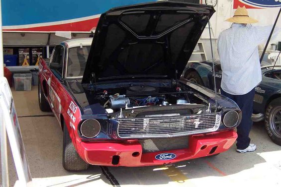 Front view of a 1965 Ford Mustang race car with hood open, red and blue paint, and racing decals. Another car is visible to the right