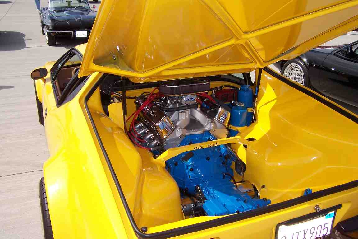 Yellow sports car with hood open showing V8 engine and blue transmission. Black car in background