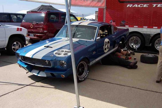 Blue 1965 Ford Mustang race car with white stripes and number 27 on door, being worked on under the hood