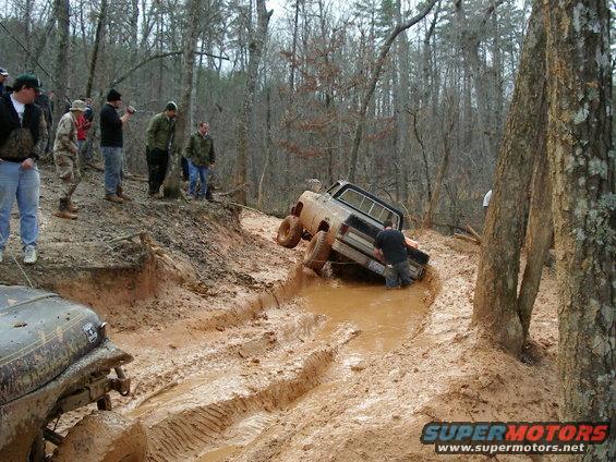 Two off-road vehicles stuck in deep mud on a wooded trail, with several people watching