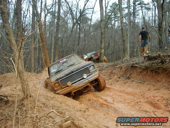 Two off-road vehicles stuck in thick mud on a wooded trail. The front vehicle is a dark-colored pickup truck