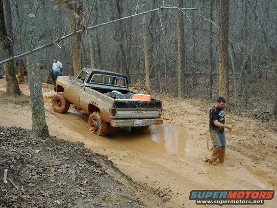 Muddy pickup truck stuck in a muddy trail with two people nearby