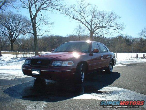 Maroon sedan parked on wet asphalt with snow in the background. The car appears to be a Ford Crown Victoria, likely from the late 1990s