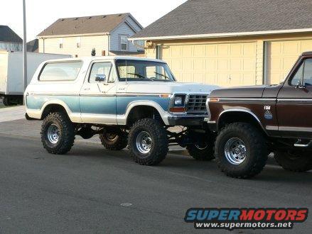Two lifted 4x4 trucks parked in a driveway. One is a white and blue 1979 Ford Bronco, the other is a brown Ford F-Series truck