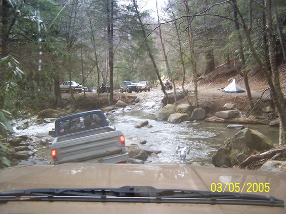 Several trucks driving through a shallow stream in a wooded area. A tent is set up on the bank
