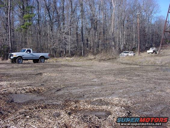 Silver pickup truck parked in a muddy area with trees behind it. Two other trucks are visible further back