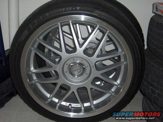 Close-up of a silver multi-spoke aftermarket wheel with a tire mounted. Some text is visible on the tire sidewall
