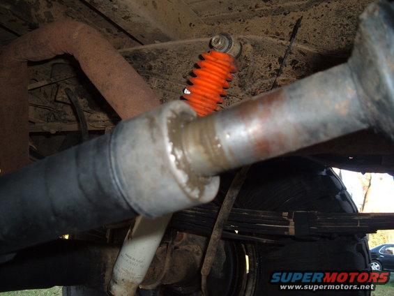 Underside of a vehicle showing exhaust pipe, orange shock absorber, and leaf springs