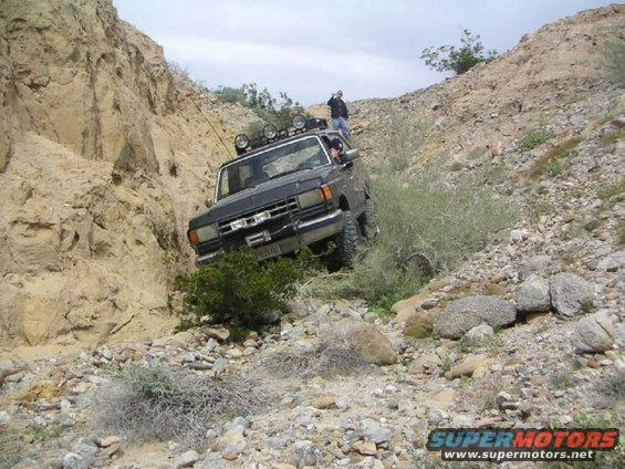 Black 1987 Ford F-150 driving up a rocky hill, with a person standing on the ridge above