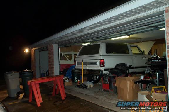 White SUV parked in a garage with another vehicle partially visible behind it. Tools and boxes are scattered around