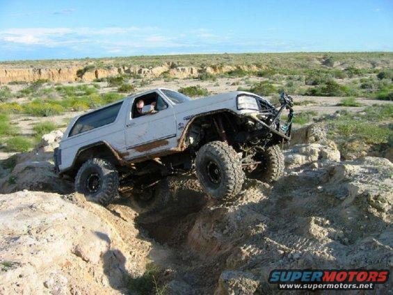 White SUV with large tires climbing rocky terrain. Modified front bumper and roll cage visible