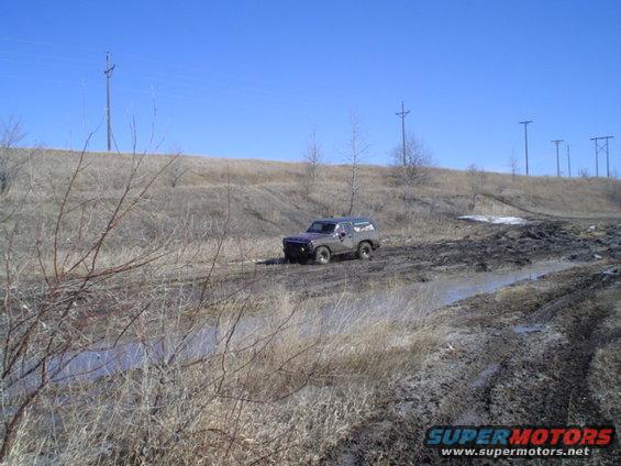 1981 Ford Bronco navigating a muddy off-road trail with dry grass and a clear blue sky