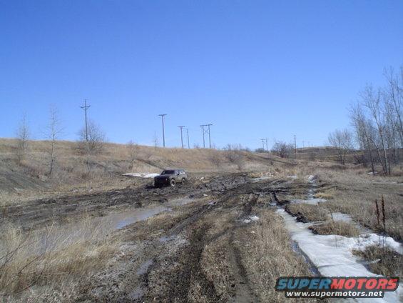 Dark SUV struggling on a muddy, rutted track with snow patches under a clear blue sky
