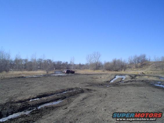 Dark colored SUV driving through a muddy, off-road area with trees and dry grass in the background under a clear blue sky