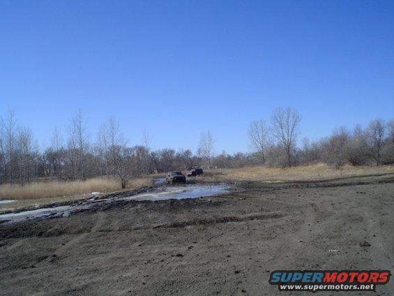 Two SUVs navigate a muddy track under a clear blue sky