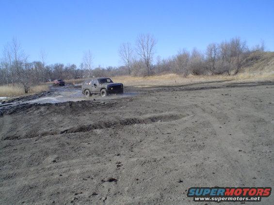 Two SUVs driving through a muddy track with trees and a clear blue sky in the background