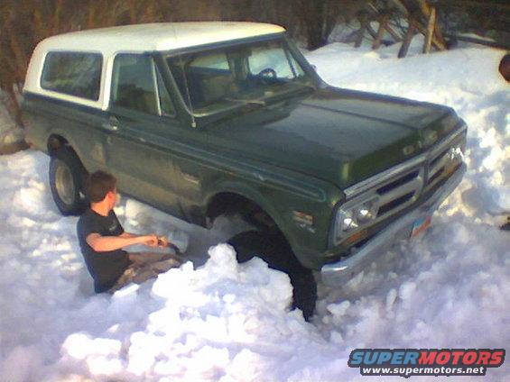 Green GMC K5 Blazer with white top stuck in deep snow, person sitting beside front wheel