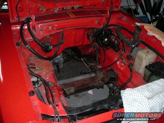 Red car engine bay with brake booster, hoses, and wiring visible. Engine is missing