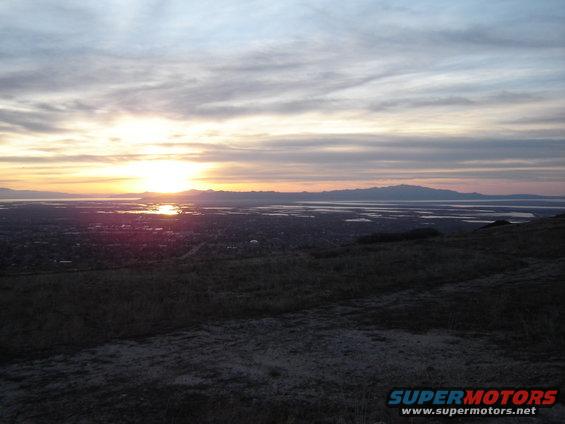Sunset over a wide body of water with mountains in the distance, viewed from a grassy hill