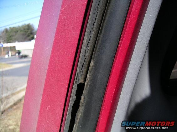 Close-up of a cracked rubber seal on a red vehicle door frame. Background shows a street and trees
