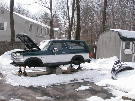 Green and white Ford Bronco on jack stands with hood open, snow on ground