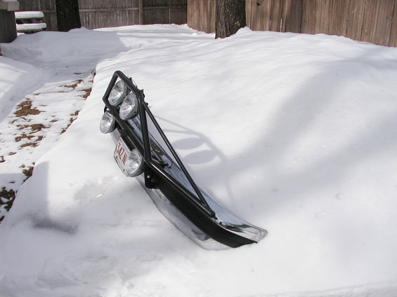 Chrome bumper with a black light bar and four auxiliary lights, partially buried in snow. A license plate is visible