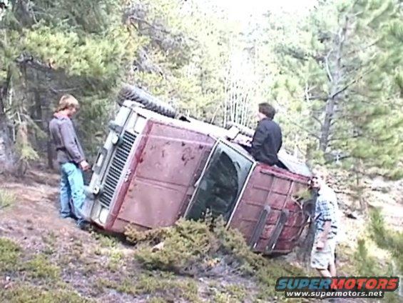 Red SUV tipped on its side in a wooded area, showing its driver's side and roof
