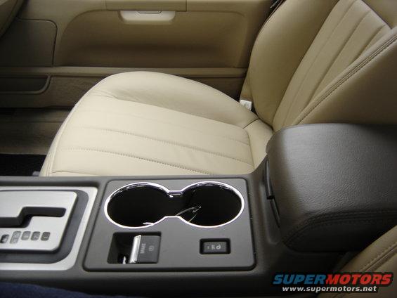 Car interior showing tan leather seats, center console with cup holders, gear shifter, and brake button. Supermotors.net logo visible