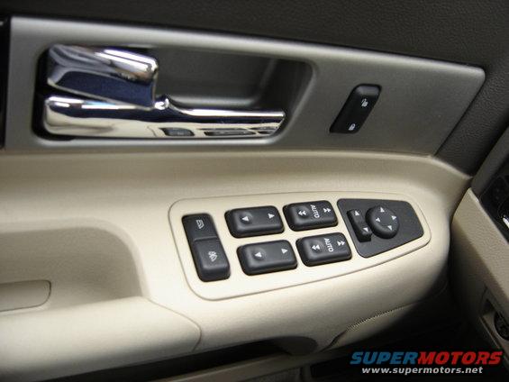 Car interior door panel with chrome handle, window controls, and mirror adjustment buttons. Beige and gray trim