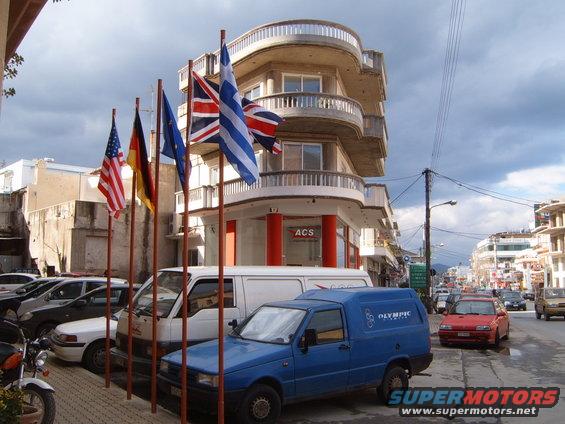 Street scene with flags of USA, Germany, EU, and UK in front of a building; parked cars and vans visible