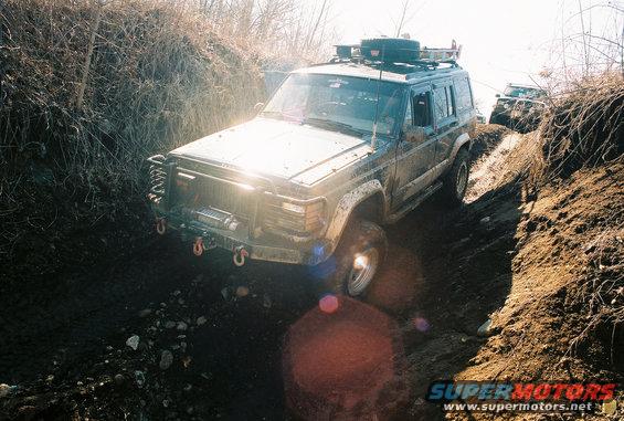 Mud-covered Jeep Cherokee XJ with a winch and roof rack driving off-road