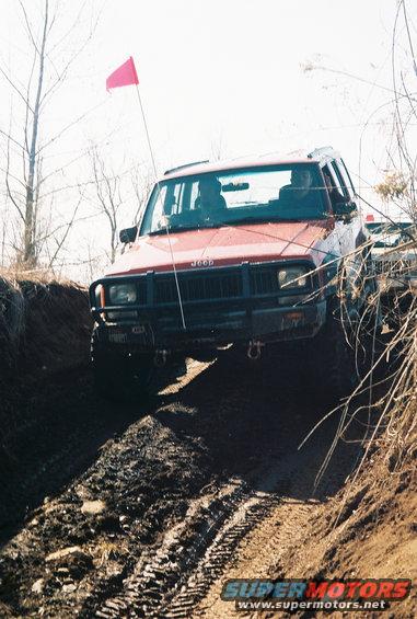 Red Jeep Cherokee with bull bar driving on a muddy trail, another vehicle visible behind it