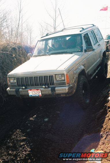 Jeep Cherokee XJ driving off-road, tires deep in mud. Another vehicle visible in background