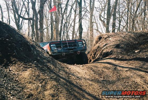 Orange off-road vehicle with bull bar driving through a dirt ditch. Red flag on a pole visible above