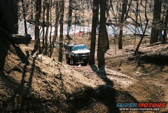 SUV driving on a dirt path in a wooded area. The vehicle appears to be a late 1990s or early 2000s model