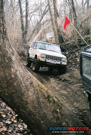 White Jeep Cherokee XJ driving on a muddy trail with trees around. Red flags on poles are visible