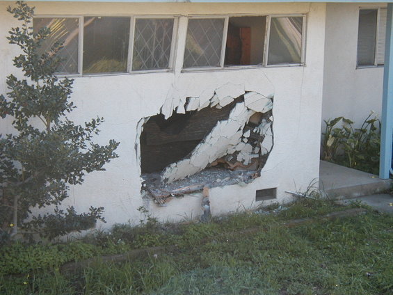 Damaged white stucco wall with large hole, exposing debris and wooden beams. Overgrown grass in foreground