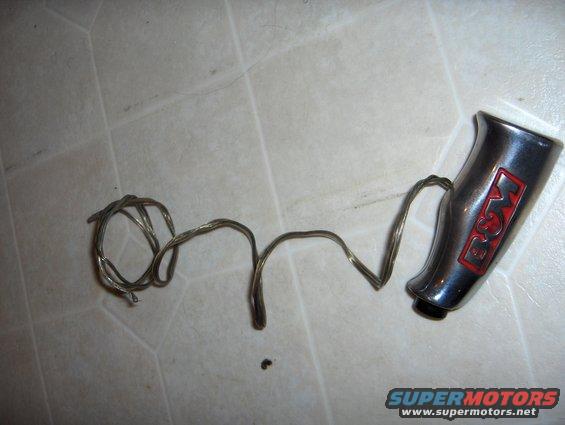 Chrome shift knob with red and white 'B&M' logo and attached wire, lying on a tiled floor