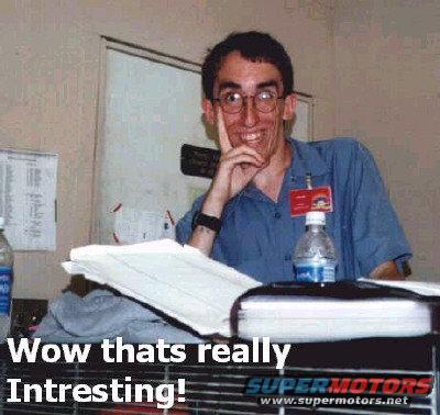 man with glasses wearing blue shirt and red badge, sitting at desk with papers and water bottle
