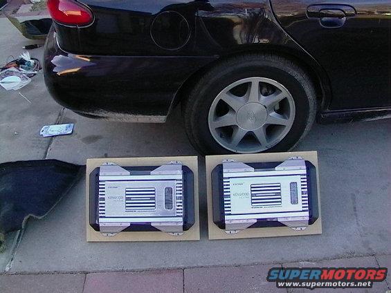 Two Kenwood car amplifiers on wooden mounts next to a black car's rear wheel