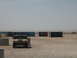 1988 Ford Bronco - A tan Humvee is parked in a lot with shipping containers in the background under a clear sky.