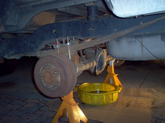 Rear axle assembly with leaf springs and brake drum, supported by jack stands. A yellow drain pan sits beneath the differential