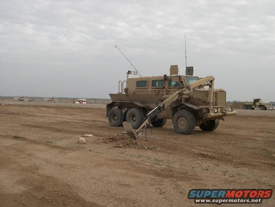 tan military truck with six wheels and an excavator arm digging in dirt