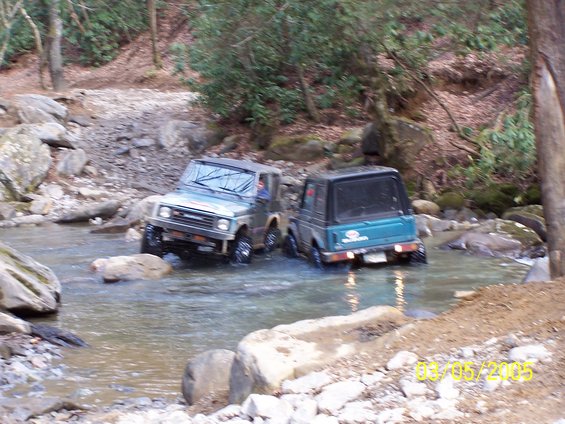 Two off-road vehicles, a light-colored SUV and a dark green SUV, drive through a rocky stream bed